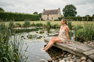 Femme assise au bord d'une piscine naturelle entourée de plantes aquatiques dans un jardin rural en France